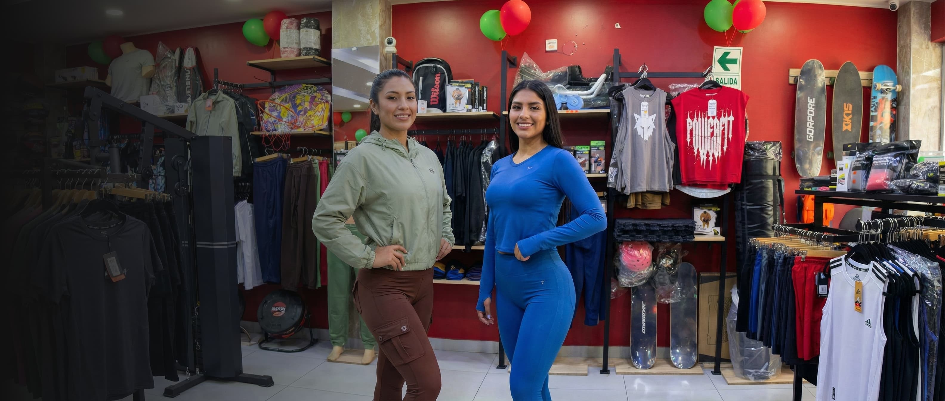 Banner de la colección de ropa deportiva para mujer en la tienda Imperium Sports. Dos modelos sonrientes posan en el interior de la tienda; a la izquierda, una mujer viste una casaca deportiva verde olivo y leggings marrones; a la derecha, una mujer luce un conjunto deportivo azul de top manga larga y leggings. De fondo se ven racks con ropa, accesorios fitness y paredes rojas.