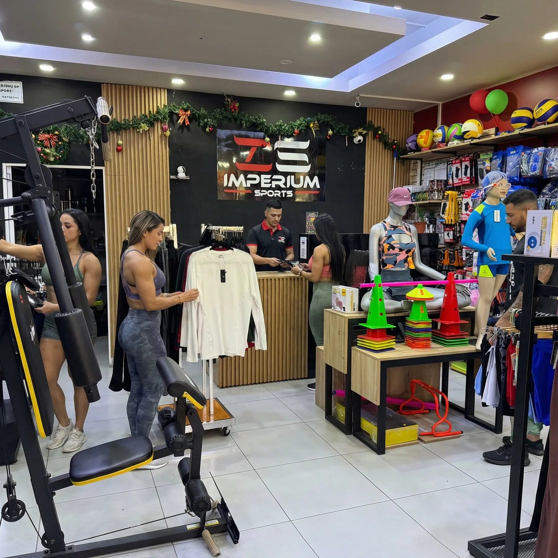 Vista amplia del interior de la tienda Imperium Sports con clientes explorando productos. A la izquierda destaca una máquina de gimnasio multifuncional; en el centro, clientes revisan ropa deportiva y leggings; y a la derecha se exhiben accesorios de entrenamiento como conos de agilidad y balones. Al fondo, el mostrador de atención con el logo de la marca y decoración navideña.