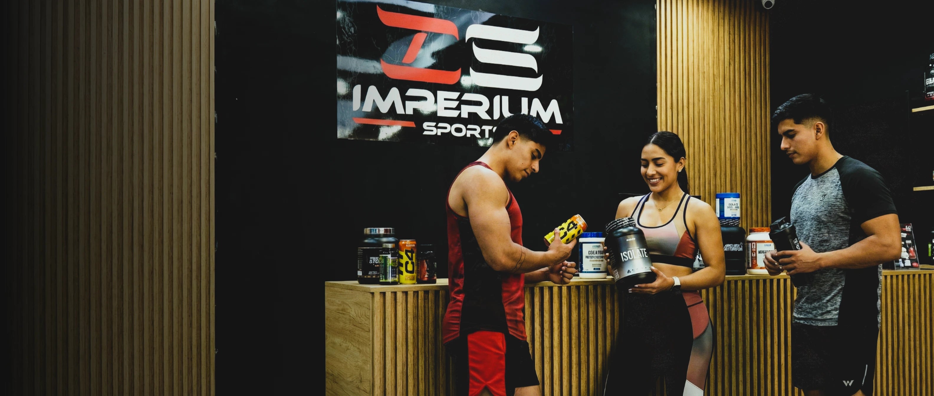 Banner del interior de la tienda de suplementos Imperium Sports, mostrando a tres atletas de cuerpo entero parados sobre el piso de concreto pulido frente al mostrador de madera. Están interactuando con productos como una proteína Isolata y una lata de pre-entreno C4 bajo el logo de la marca.